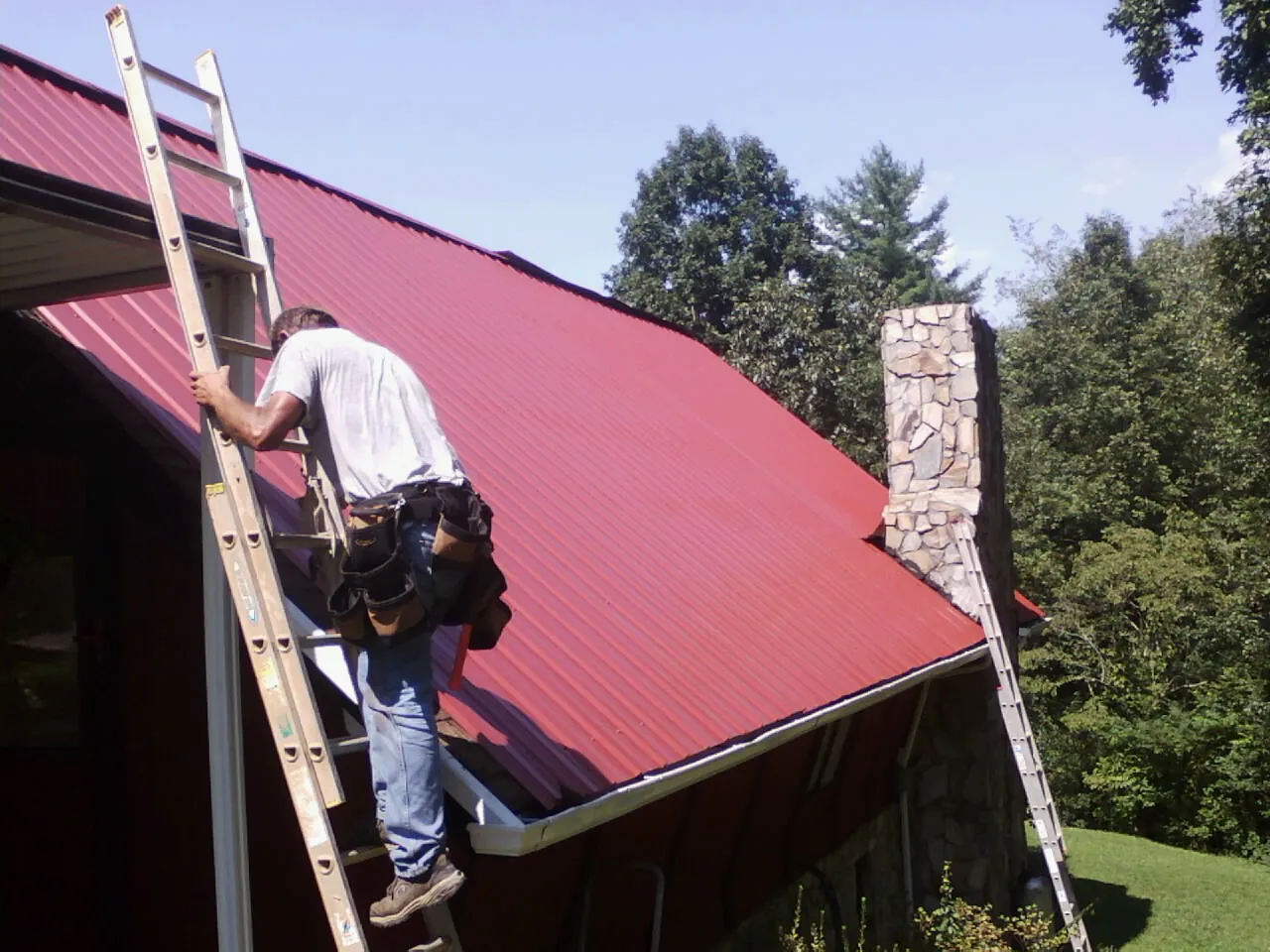 Roofer on a ladder beside a red metal roof and stone chimney — Soffit & Fascia Repair by Roofing Brothers in New York